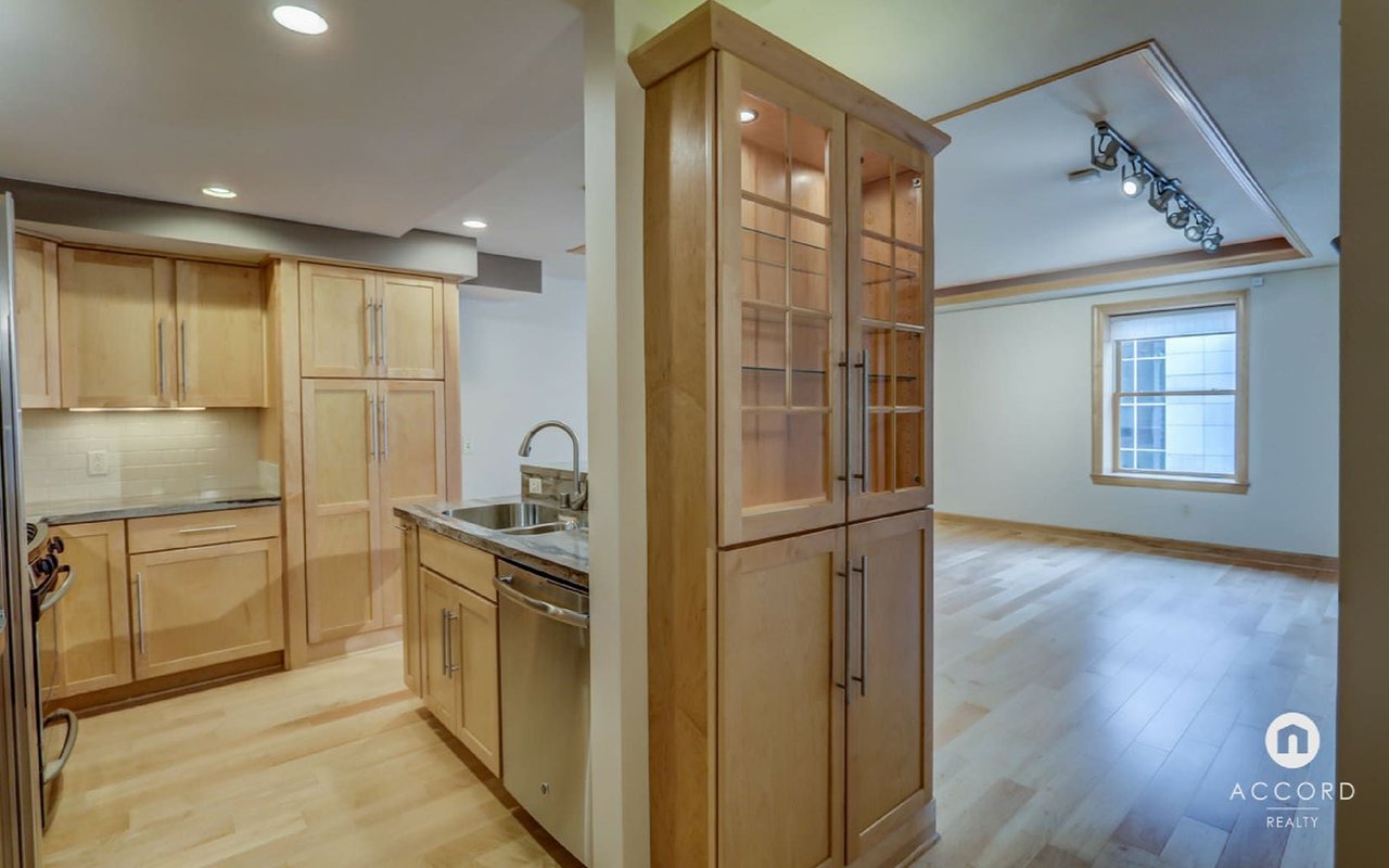 Open-plan kitchen with light wood cabinets and an adjoining empty living area.