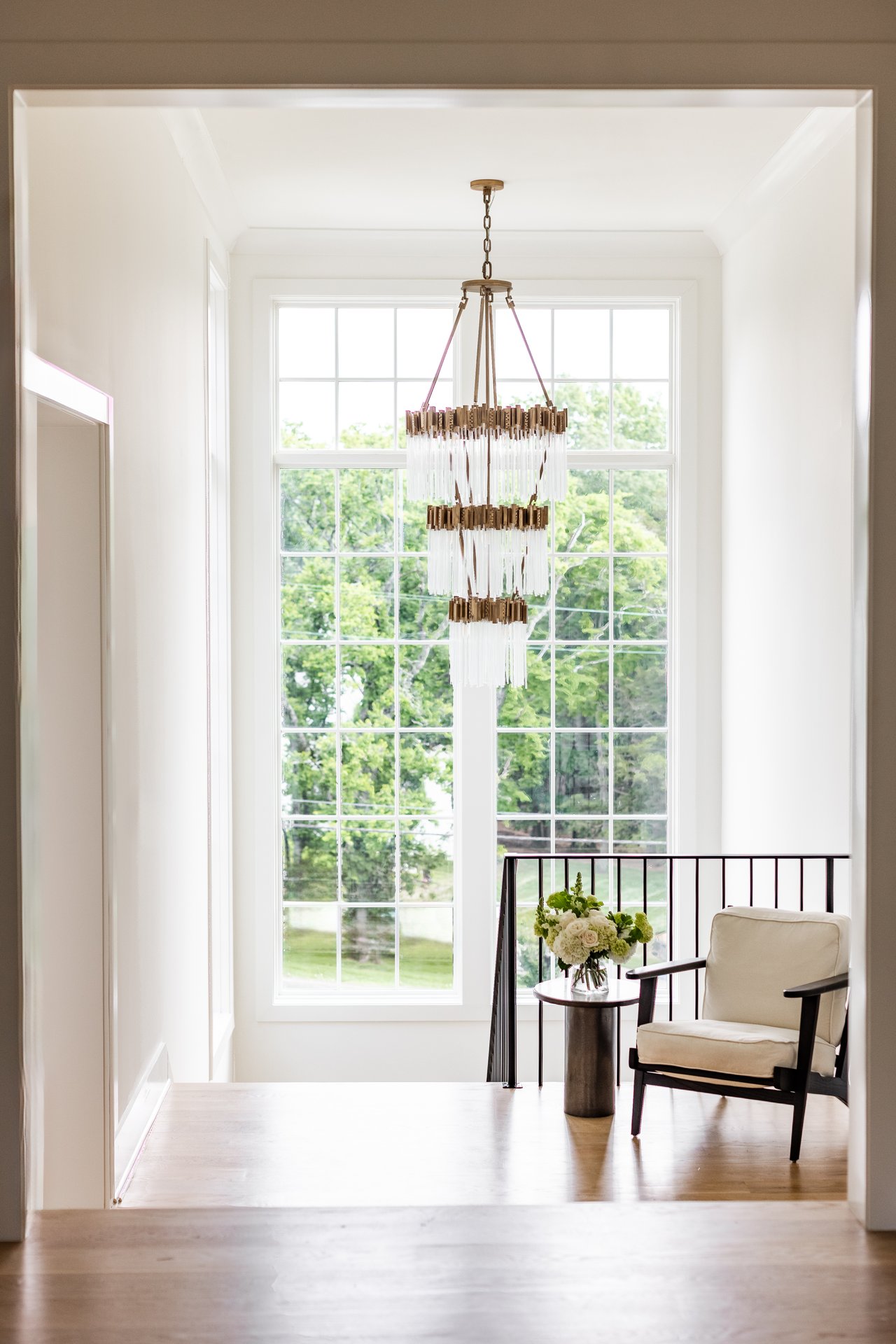 Bright sitting area with a tiered chandelier, tall windows, and a cream chair beside a flower arrangement.