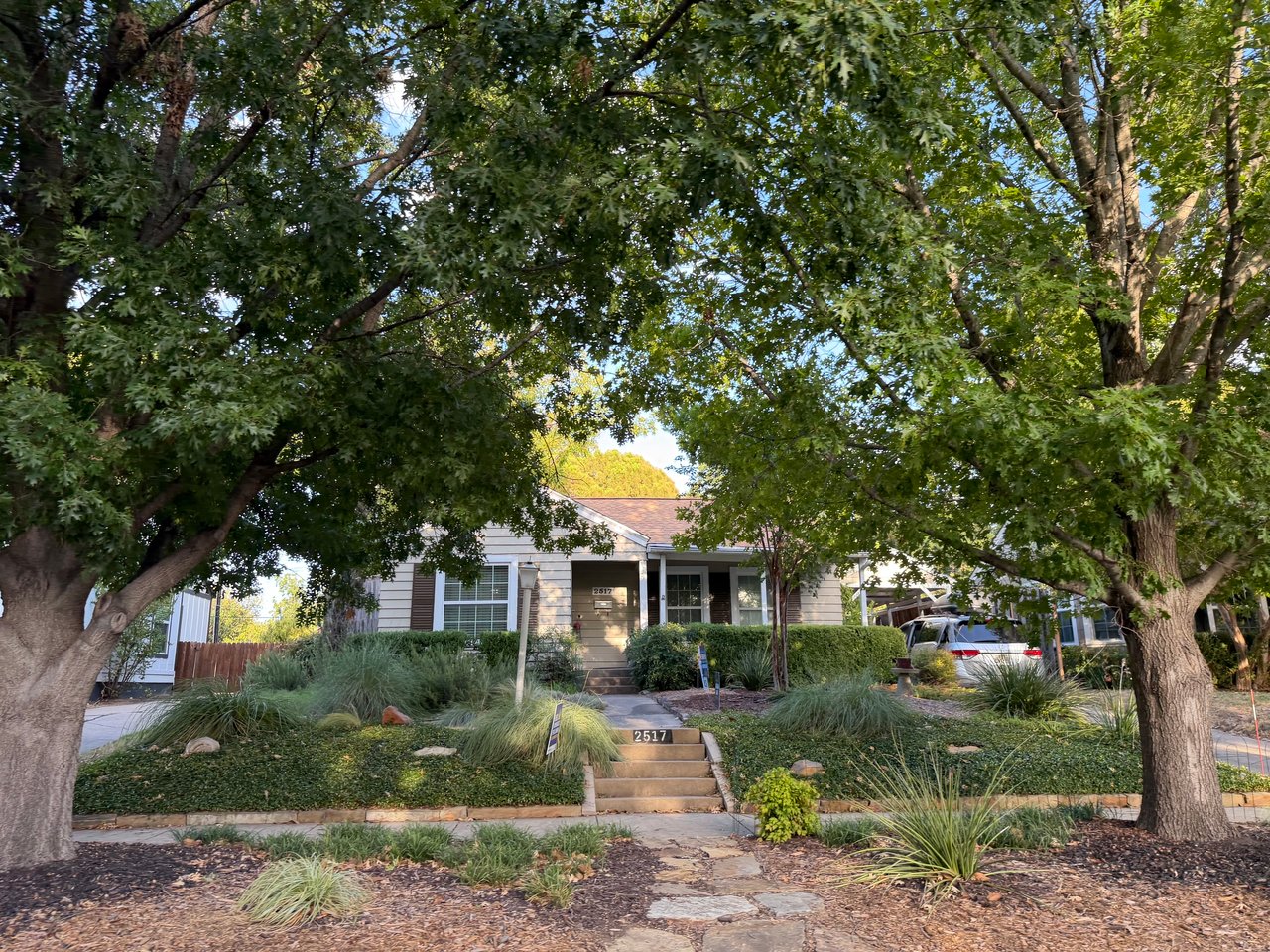 Single-story home behind a landscaped front yard, framed by large mature trees.