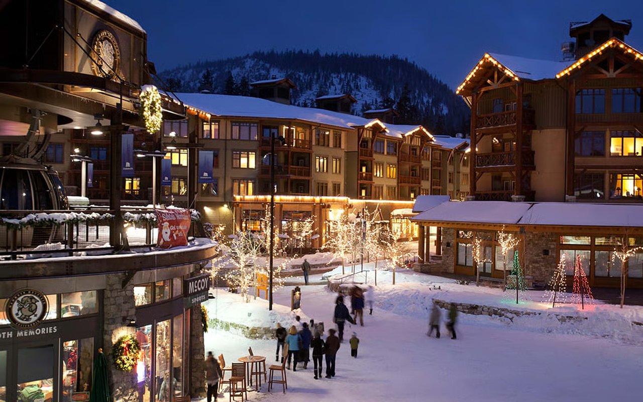 Snowy mountain resort plaza at night, with illuminated lodge buildings and holiday lights.