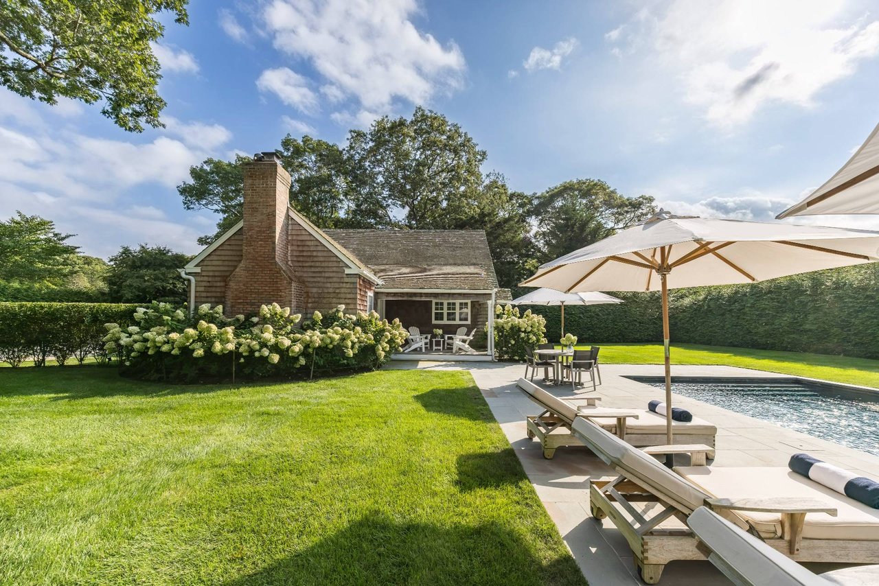 Shingled cottage beside a pool, with hydrangeas, patio umbrellas, and outdoor lounge chairs.