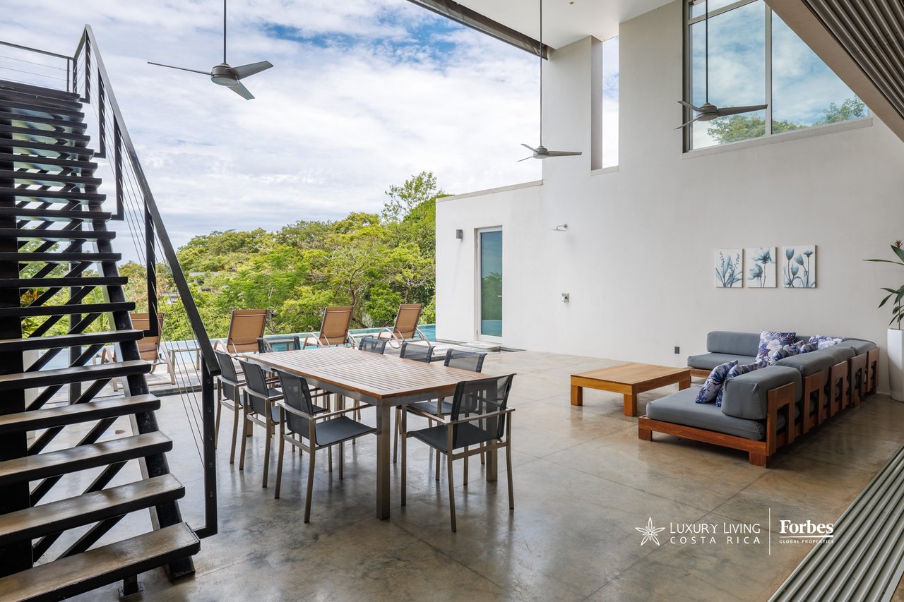 Open-plan living and dining room with a black staircase, tall ceilings, and large windows overlooking a pool.