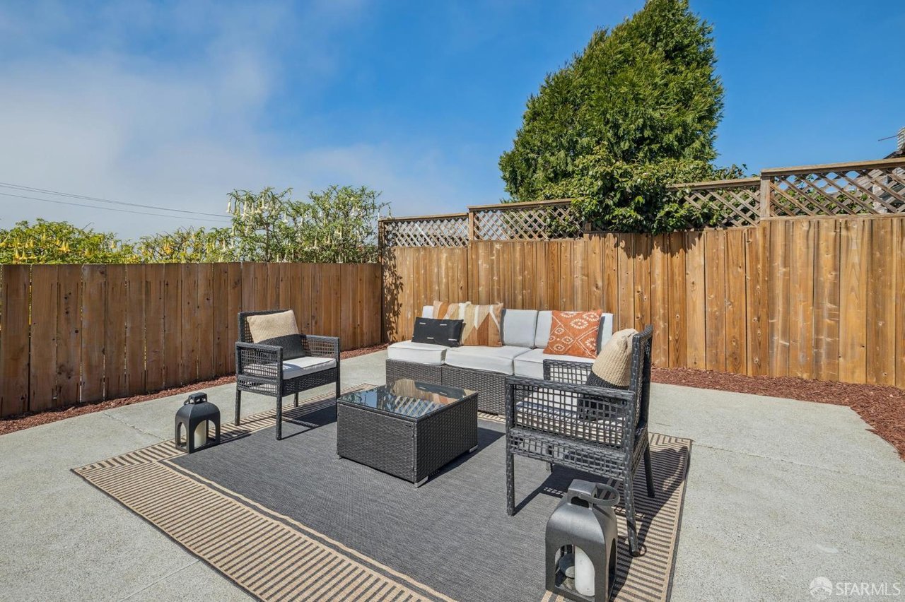 Backyard patio with outdoor sofa and chairs on a rug, enclosed by a tall wooden privacy fence.