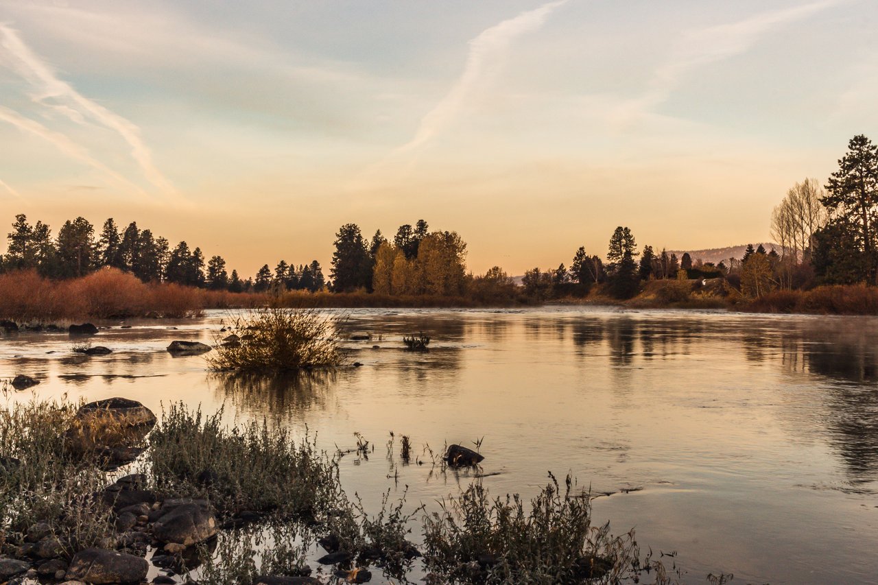 River reflecting a golden sunset, with rocky banks, brush, and autumn trees.