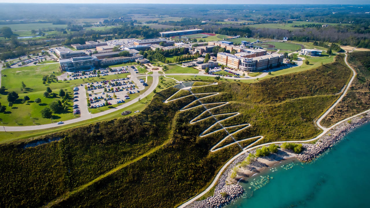 Aerial view of a large Mequon campus with parking lots, buildings, walking paths, and Lake Michigan shoreline.