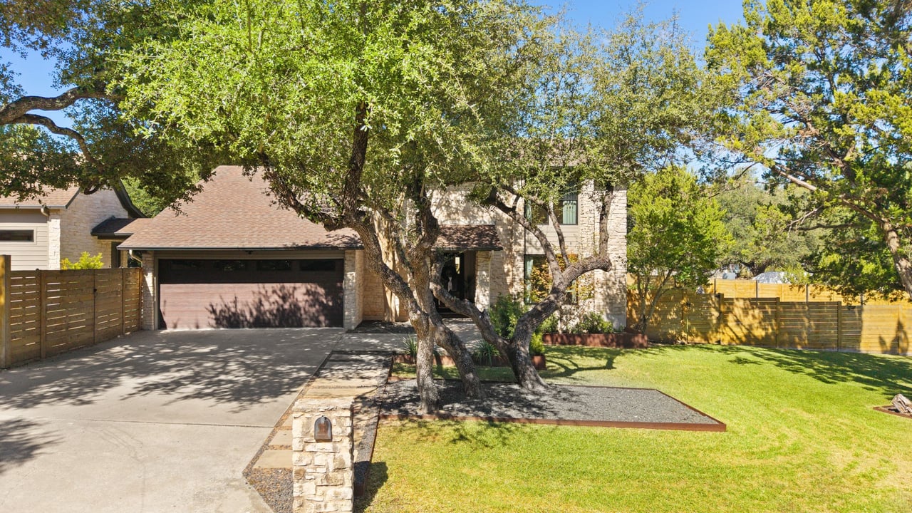Two-story stone home with a wide garage, mature shade tree, and fenced front lawn.