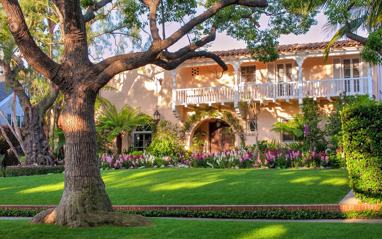 Spanish-style home with a white balcony and garden beneath a sprawling oak tree.