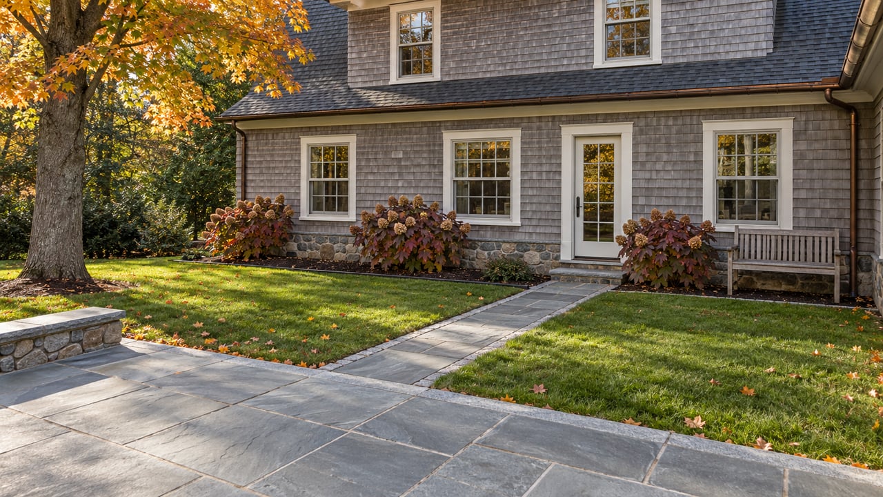 Weathered cedar-shingle home beside a bluestone terrace and autumn lawn, framed by an orange-leaved sugar maple.