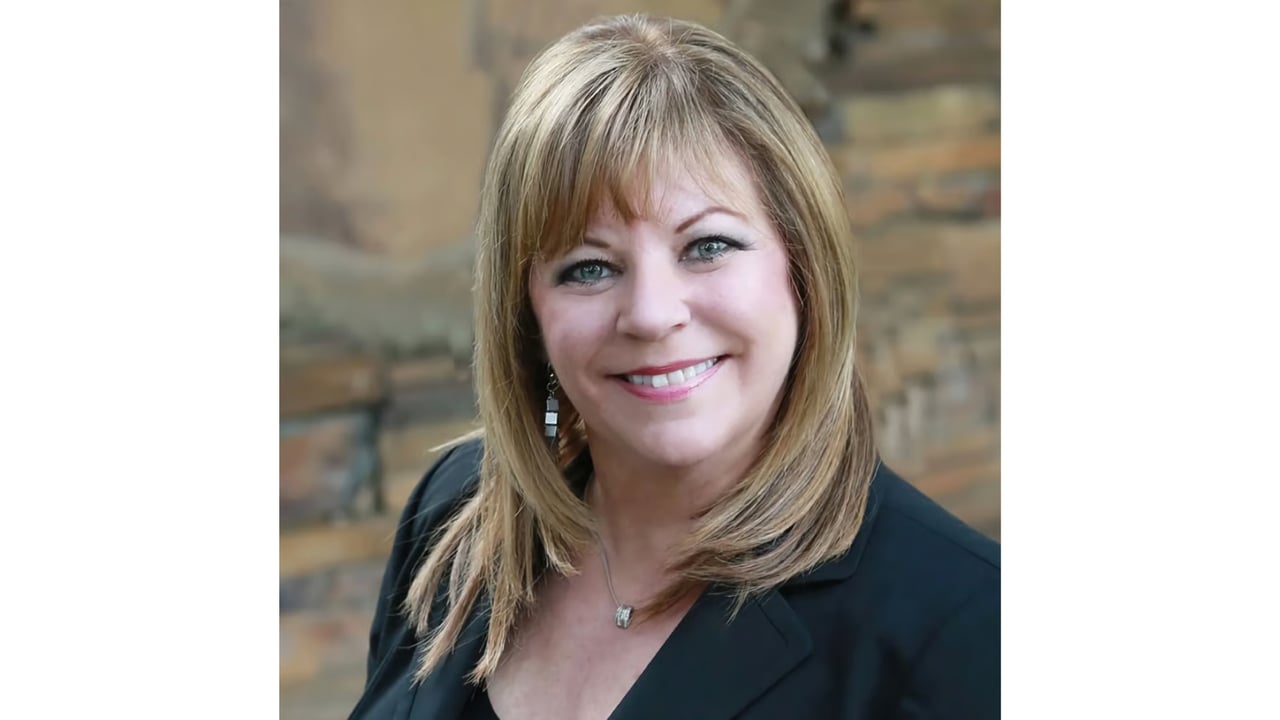 Linda Traurig smiling in a black blazer against a brick wall.
