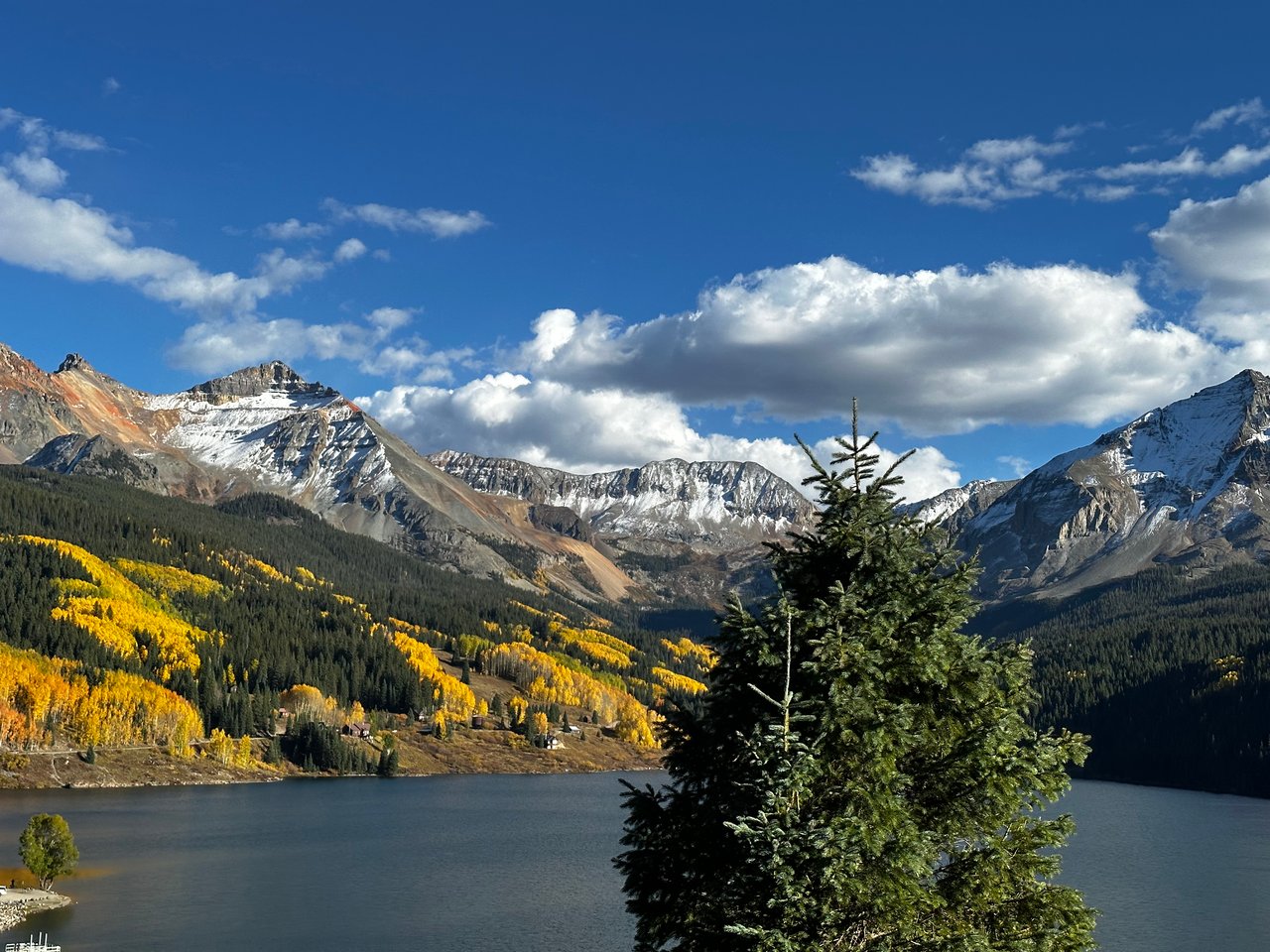 Snow-dusted mountains and golden autumn trees surround a lake, with an evergreen in the foreground.