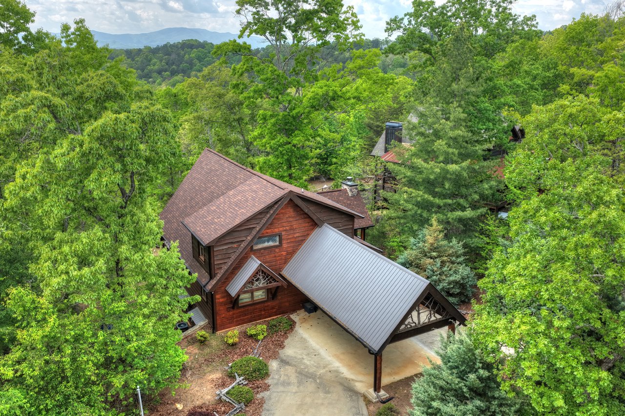 Aerial view of a wood-sided mountain home among trees, with a metal-roofed carport and distant mountain ridges.