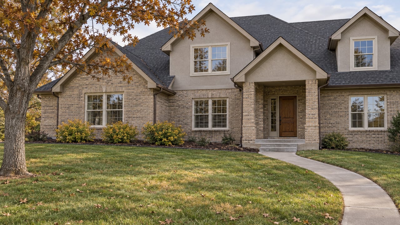 A substantial gabled brick-and-stucco home behind a broad autumn lawn, with a curved concrete approach and a mature oak.