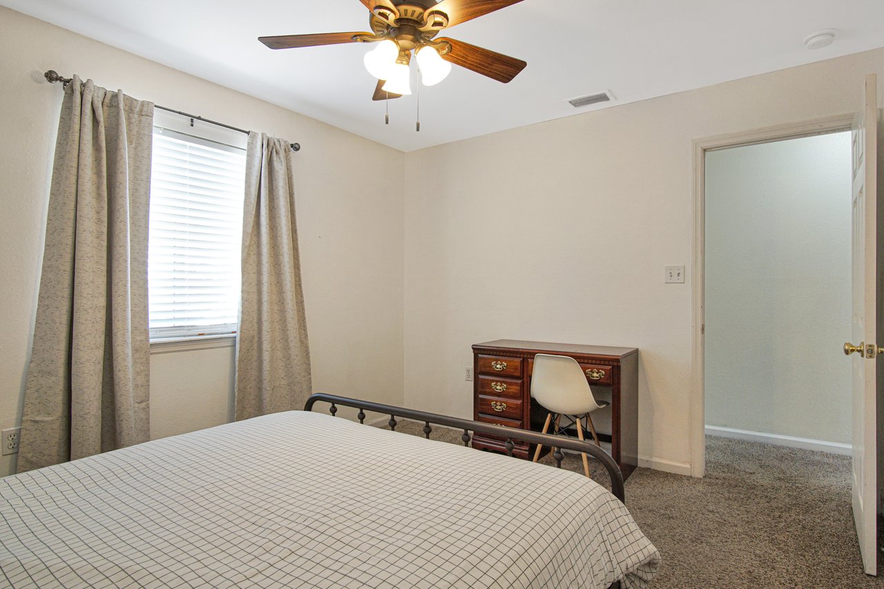 Cozy bedroom with a checkered bedspread, wooden desk with a white chair, beige curtains over a window, ceiling fan, and open door, conveying warmth.
