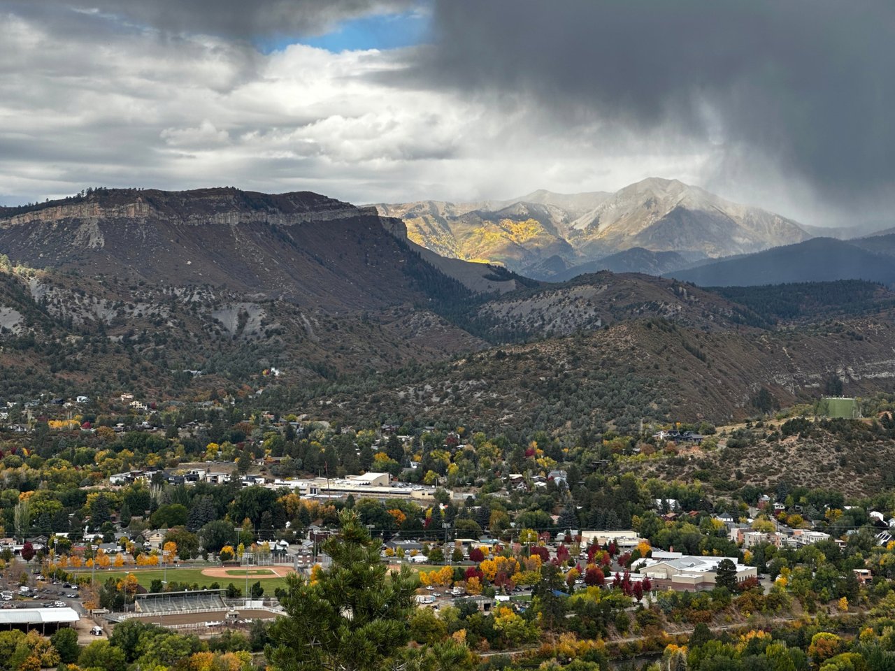 Mountain town and autumn-colored foothills beneath distant peaks and dramatic clouds.