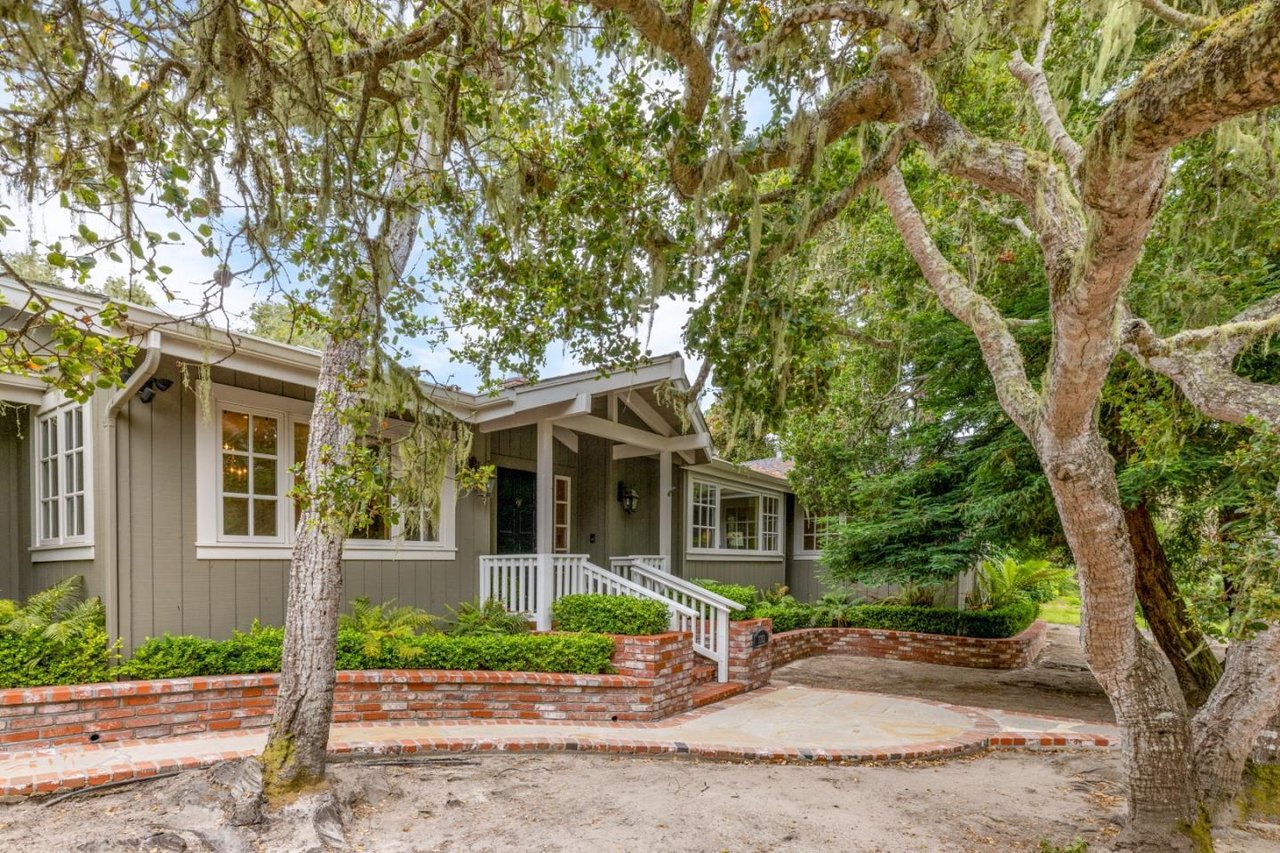 Gray home with a covered entry and curved brick garden wall beneath mature trees.