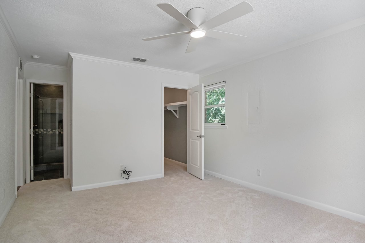 Empty room with beige carpet, white walls, and a ceiling fan. An open door leads to a closet; a small window shows greenery outside, creating a calm atmosphere.