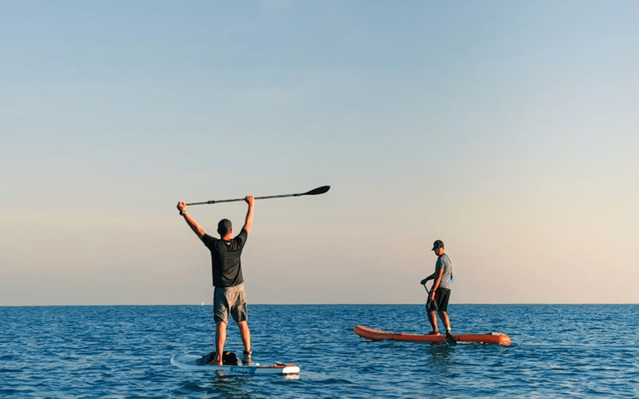 Two people paddleboard on open water, one raising a paddle overhead in warm evening light.