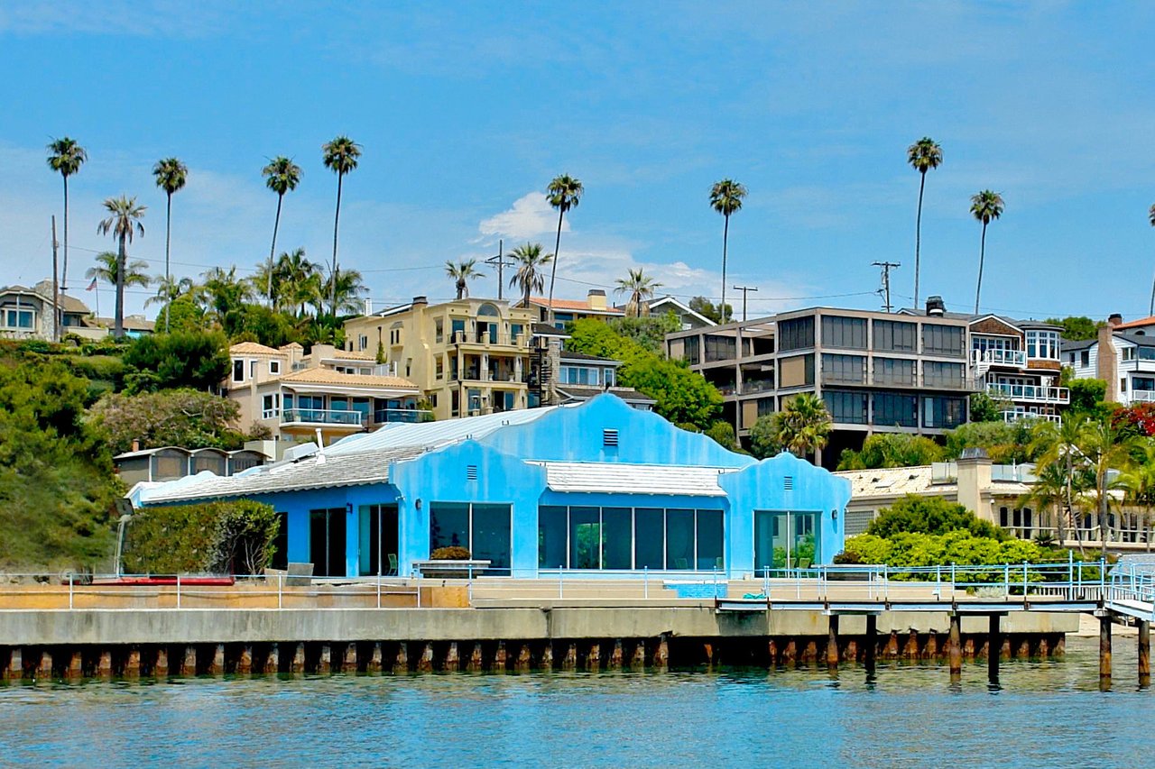 Bright blue waterfront home beside a pier, with hillside houses and palm trees behind it.