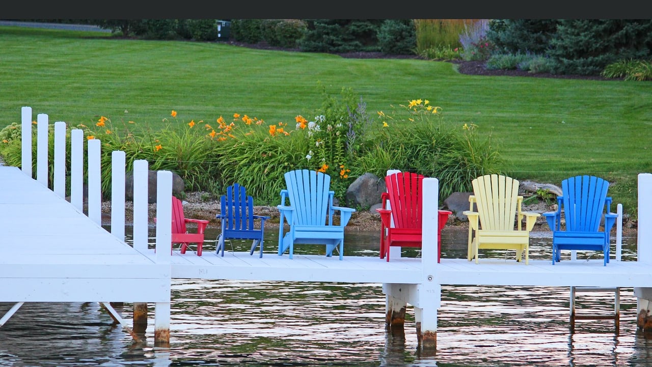 White dock over the water lined with colorful Adirondack chairs beside a landscaped lawn.