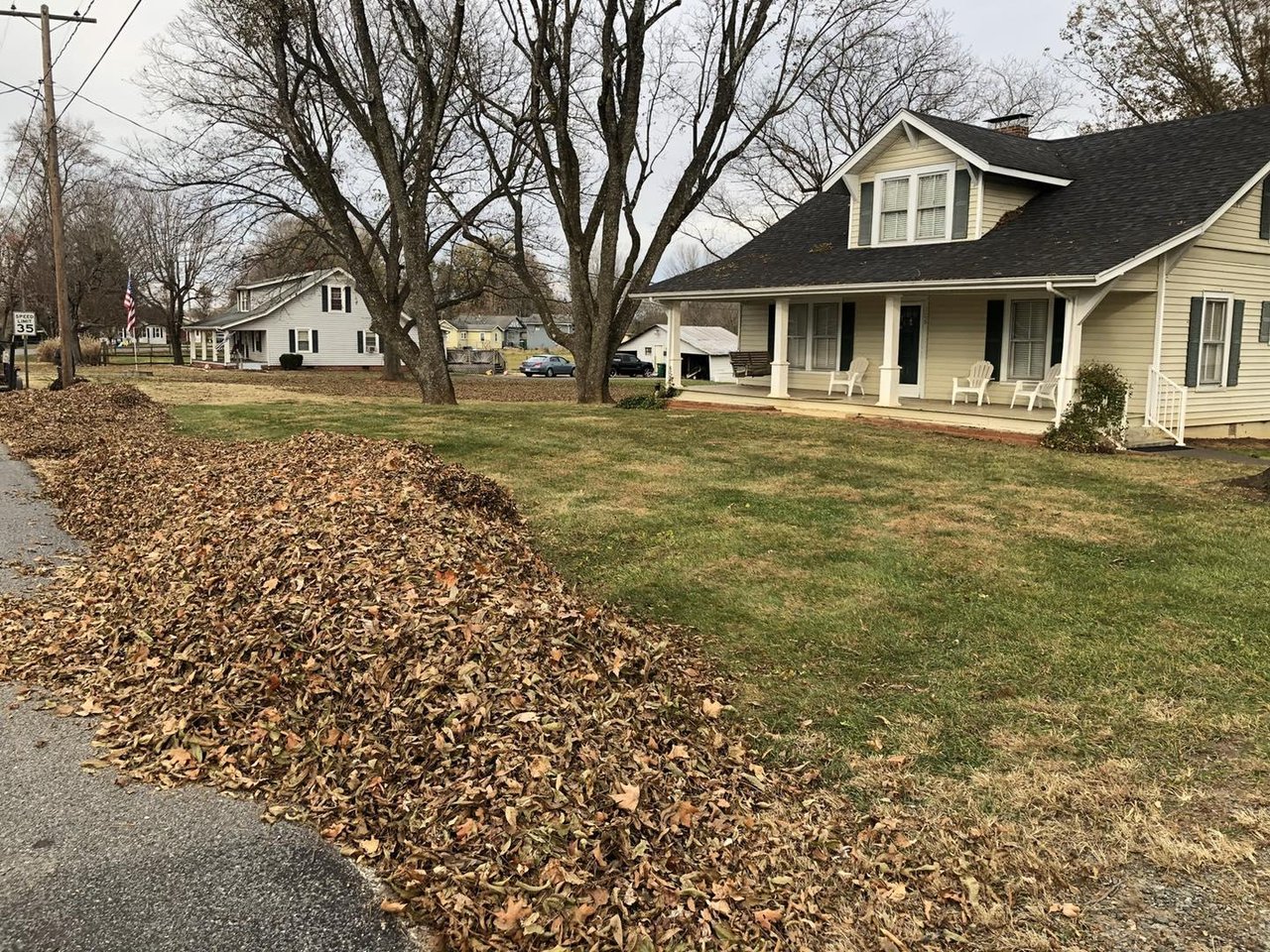Light-colored home with a covered porch and chairs beside a lawn and piles of autumn leaves.