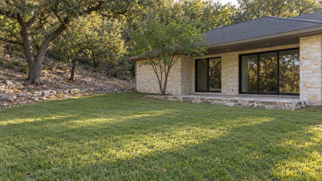 Pale limestone house beside a sloping lawn that meets a wooded rise of mature oak trees.