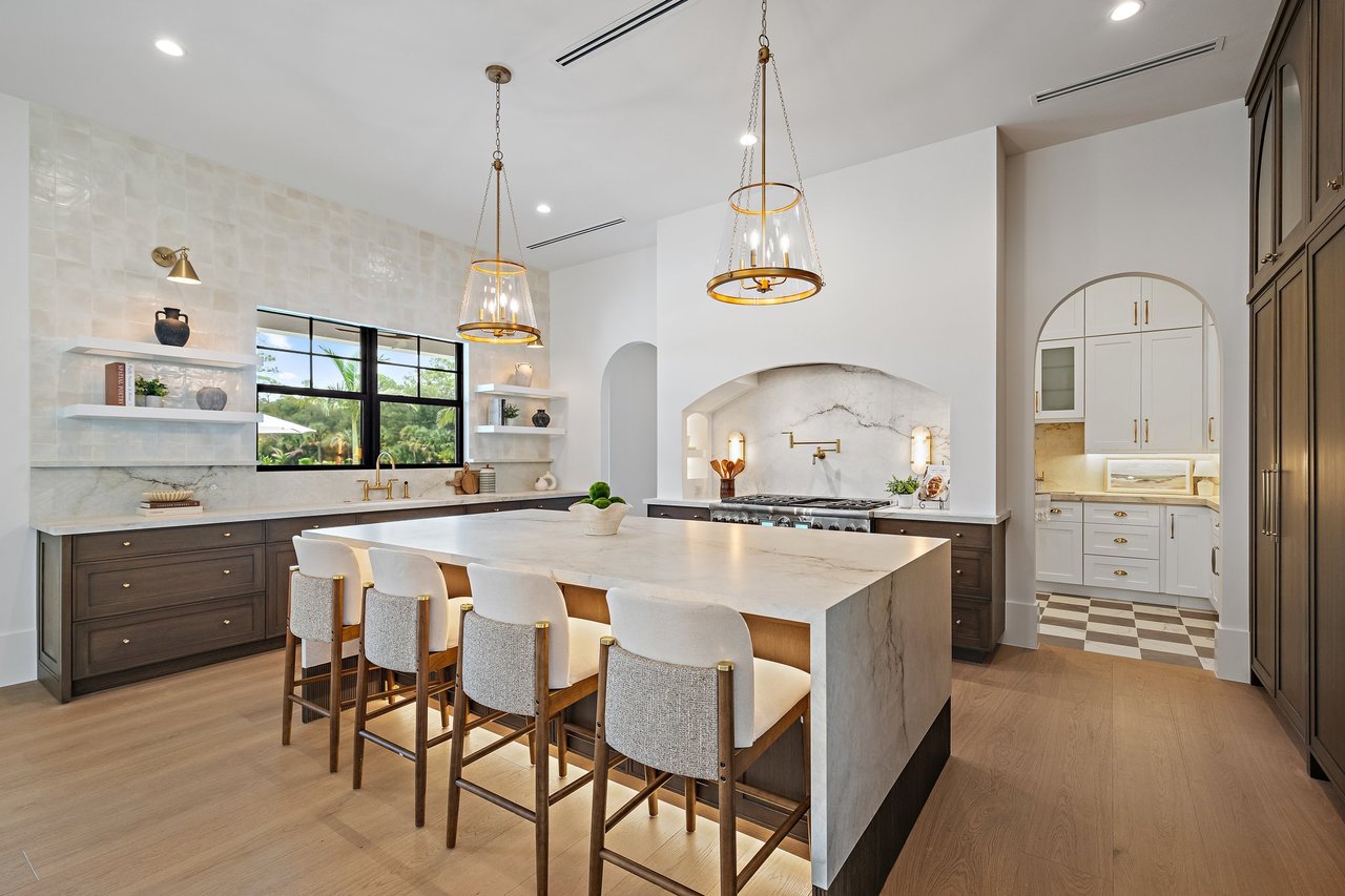 Luxury kitchen with a marble island, brass pendants, dark cabinetry, and an arched range alcove.