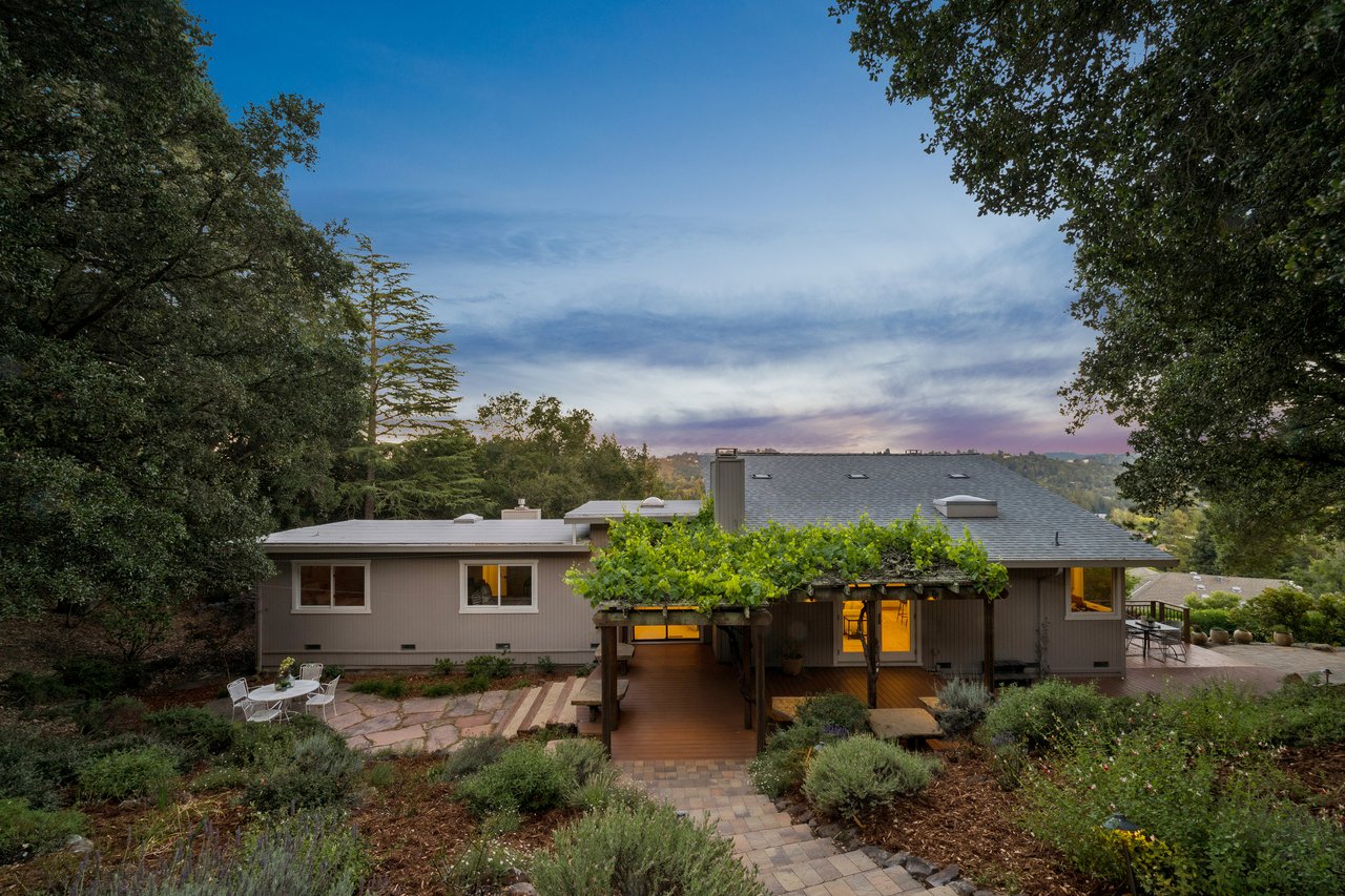 Single-story home with a vine-covered pergola, deck, and landscaped garden at dusk.