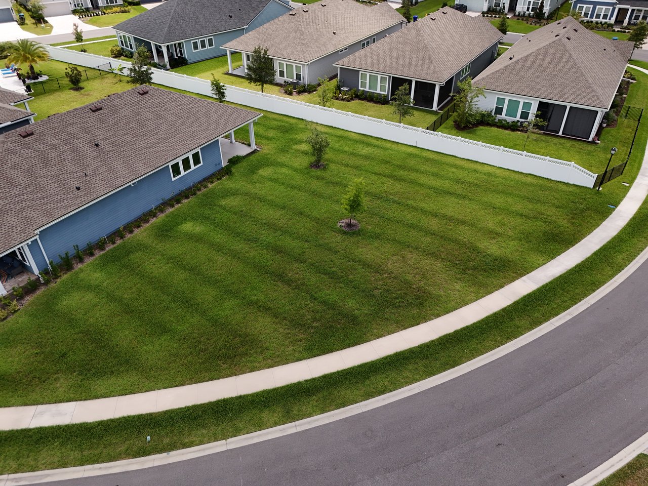 Aerial view of a grassy corner lot bordered by homes, white fences, and a curved sidewalk.
