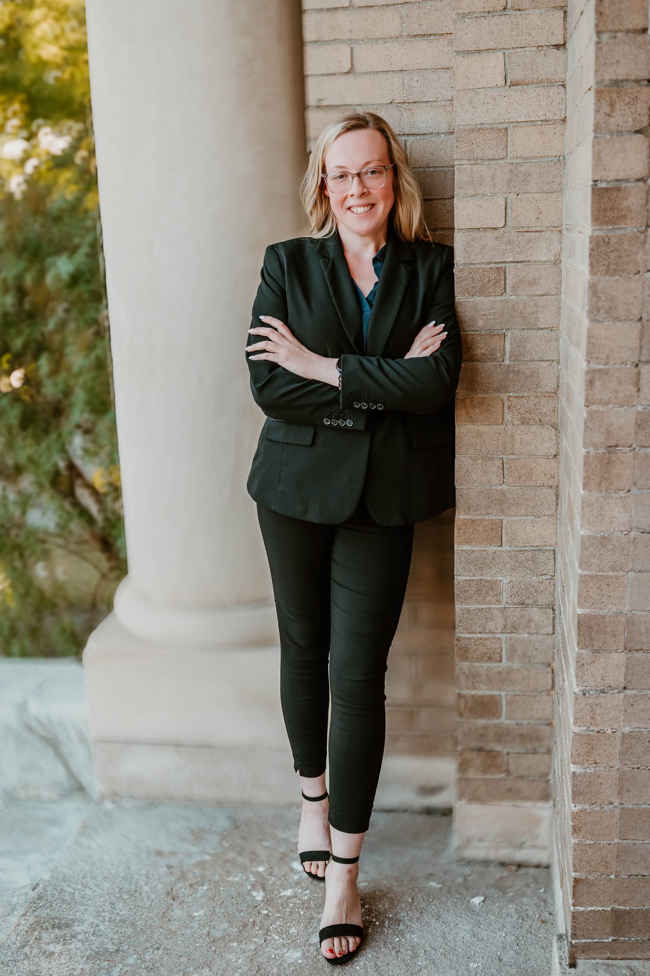 Kaeli Wager in glasses and a black suit, standing with arms crossed beside a brick column.