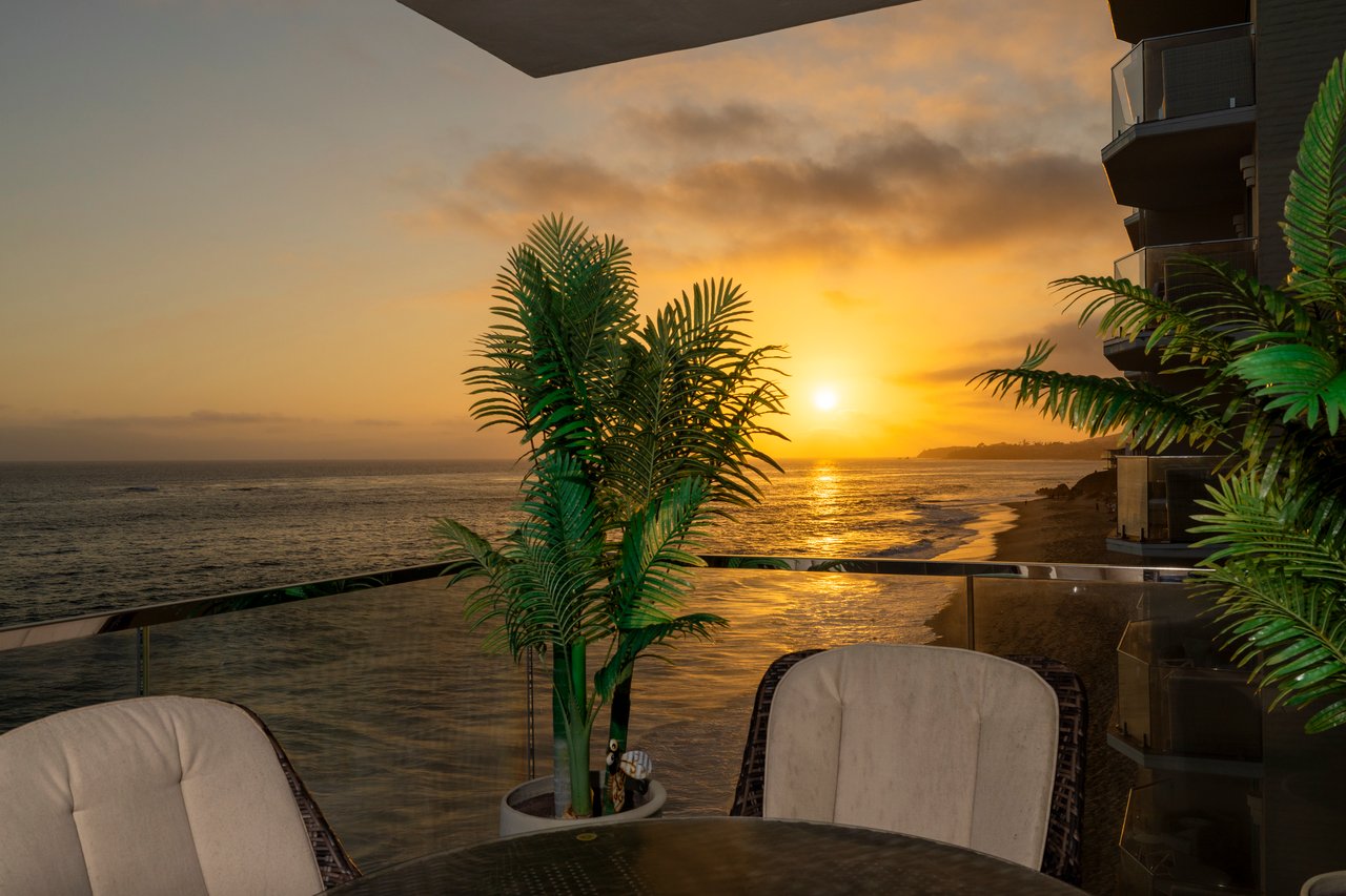 Oceanfront balcony with tropical palms and outdoor seating overlooking the sunset-lit water.