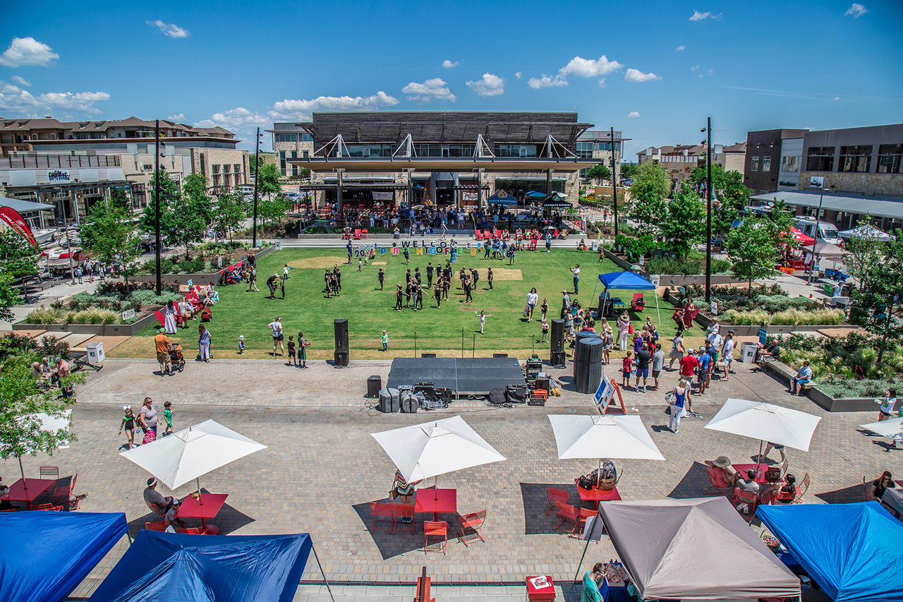 Bee Cave plaza with a community event on the lawn and shops surrounding the gathering space.