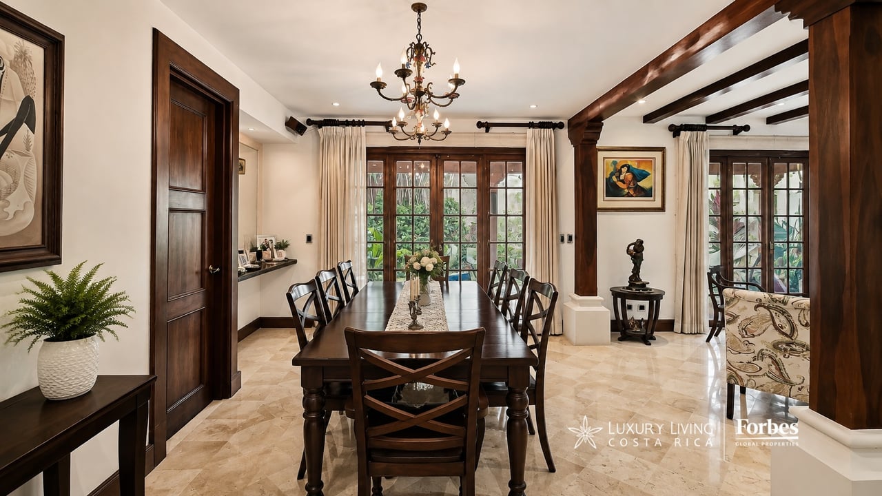 Dining room with a long dark wood table, chandelier, and French doors overlooking greenery.