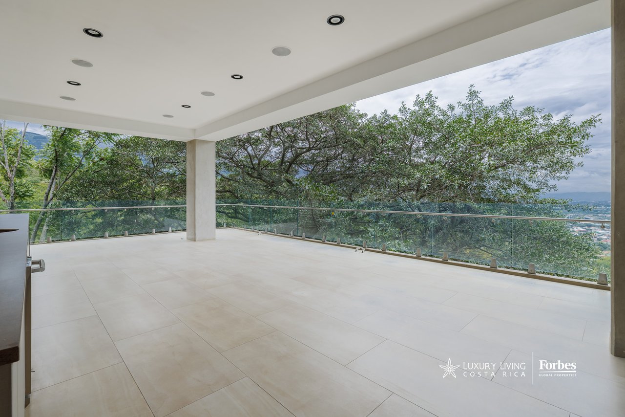 Covered apartment terrace with pale tile flooring, glass railing, and views of trees, hills, and the city.