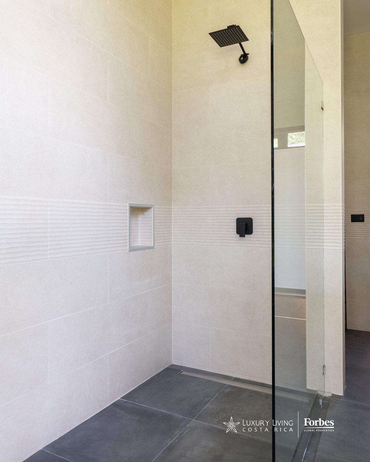 Walk-in shower with pale tiled walls, a black rainfall showerhead, and a clear glass partition.