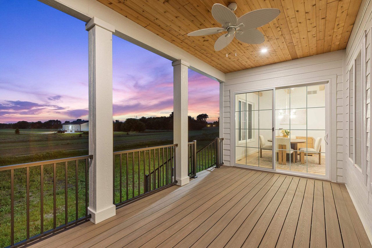 A clean, well-maintained covered composite deck with a wood plank ceiling and modern ceiling fan, looking into a staged dining room through sliding glass doors. The inviting outdoor space overlooks a green field at sunset, highlighting the importance of exterior home prep and clean landscaping.