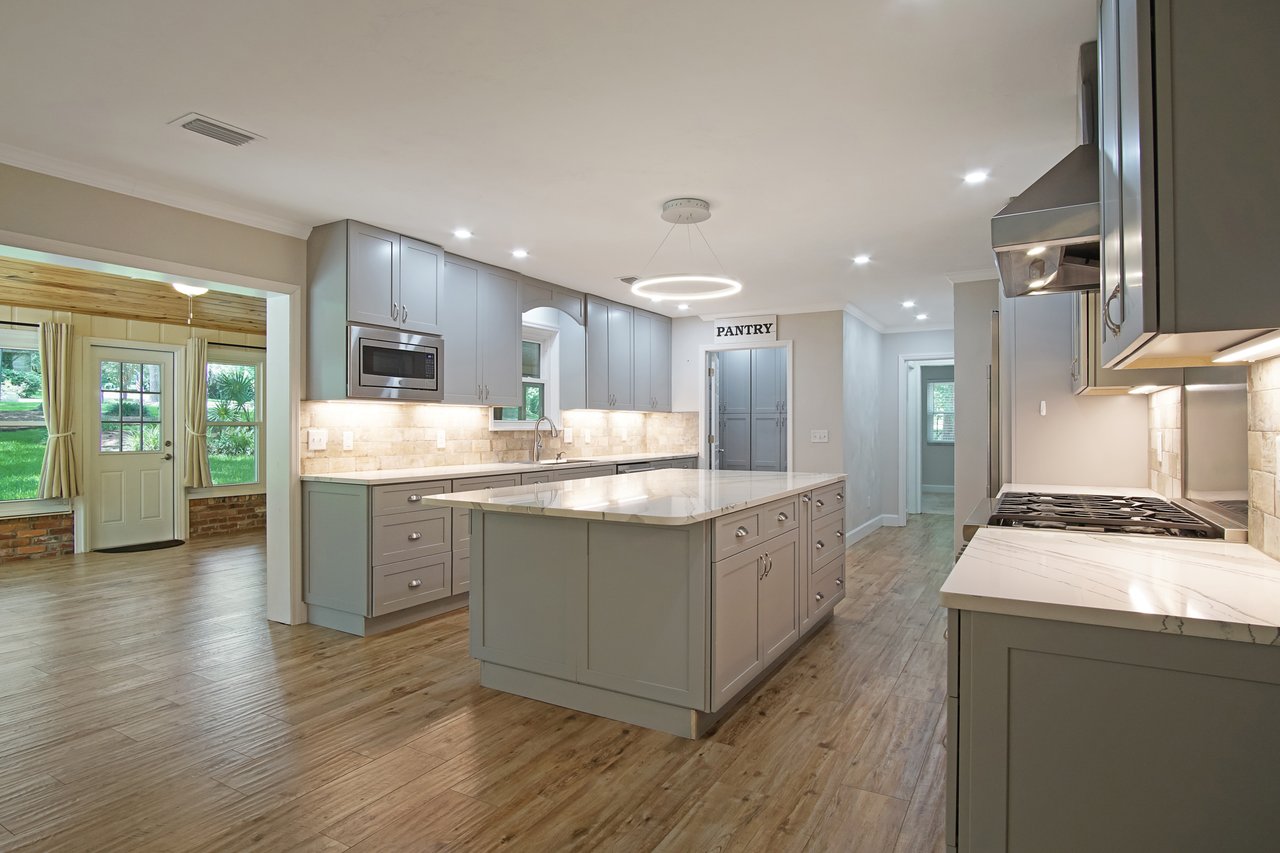Spacious kitchen with gray cabinets, large island, and wood flooring. Modern appliances, bright lighting, and a pantry door create a welcoming feel.