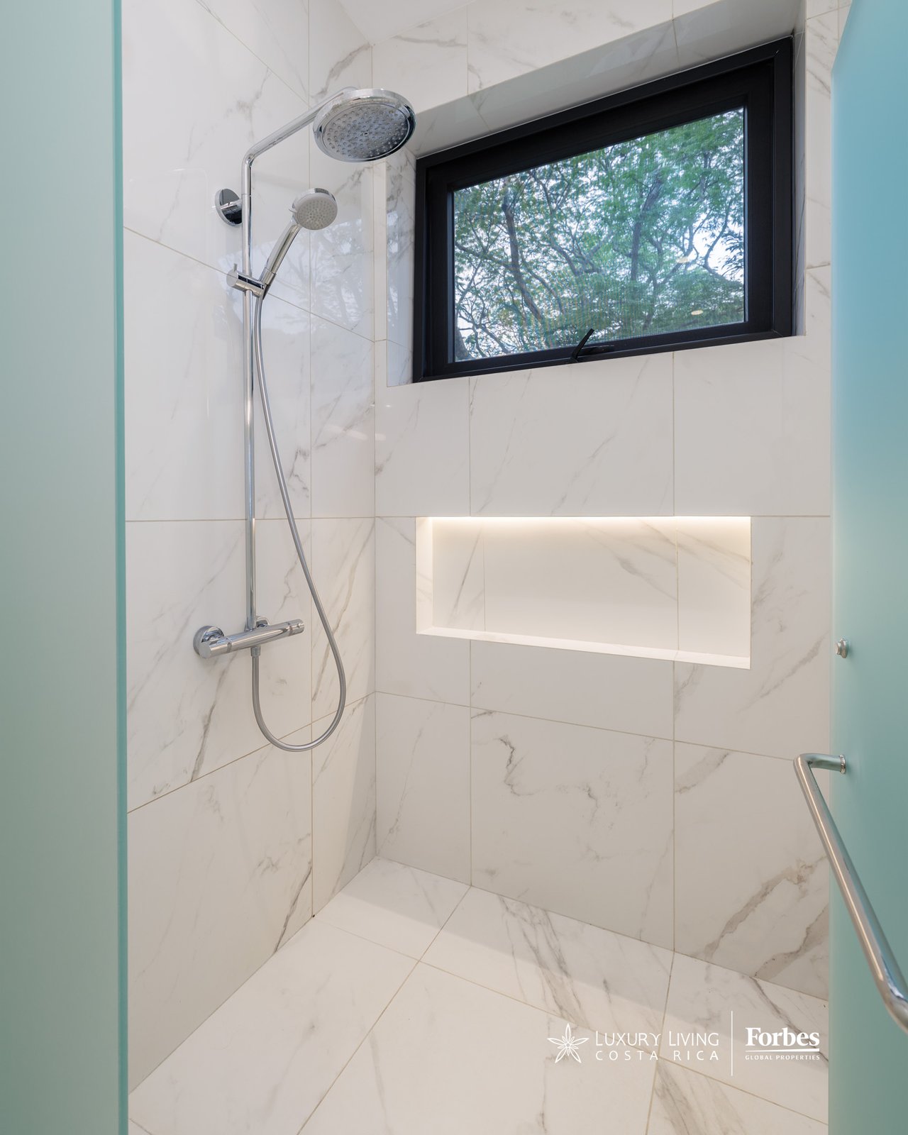 Marble-tiled walk-in shower with a rainfall head, lit niche, and window overlooking trees.