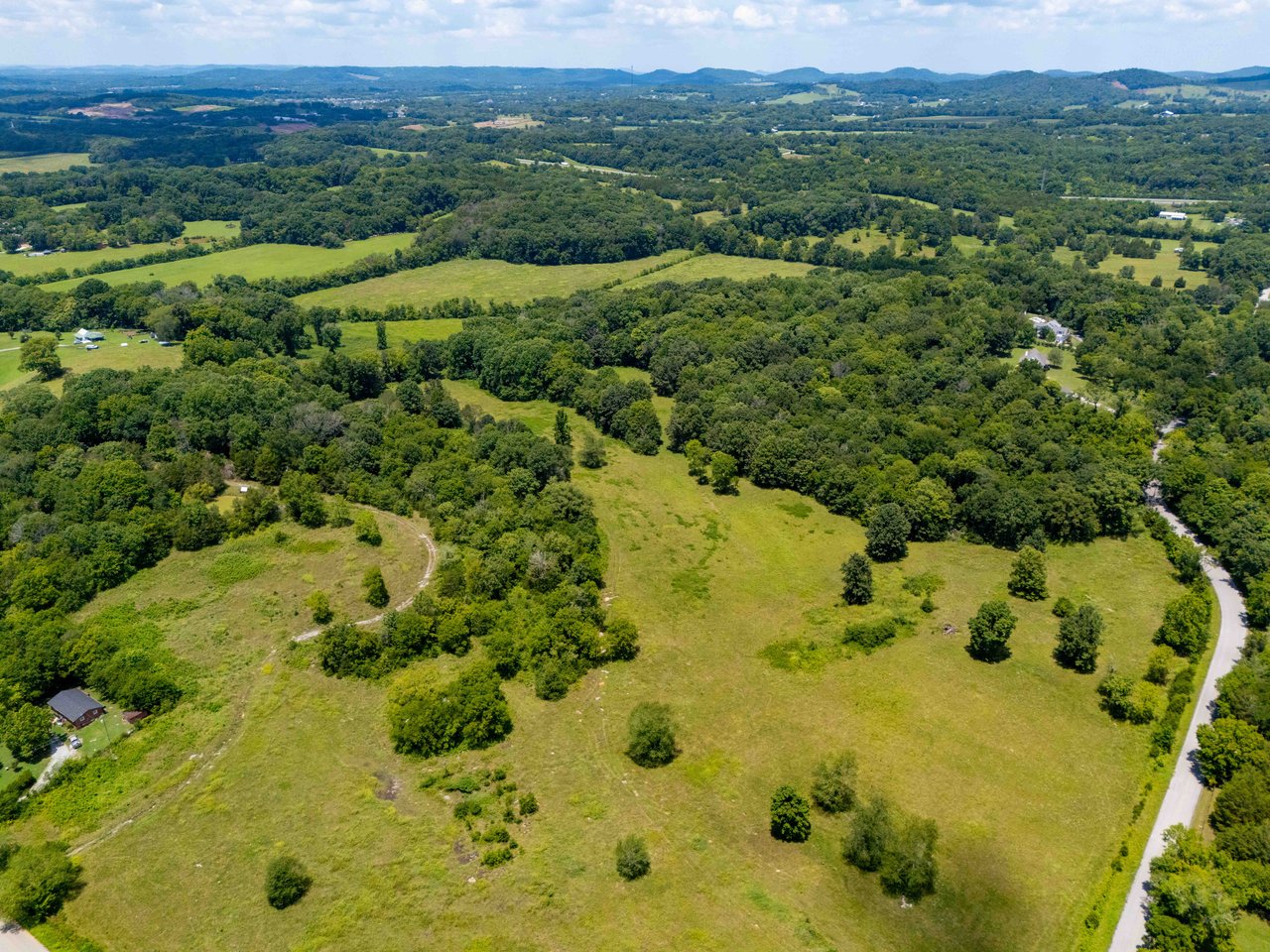 Aerial view of open grassy acreage bordered by woods, with a road along one side and rolling hills beyond.