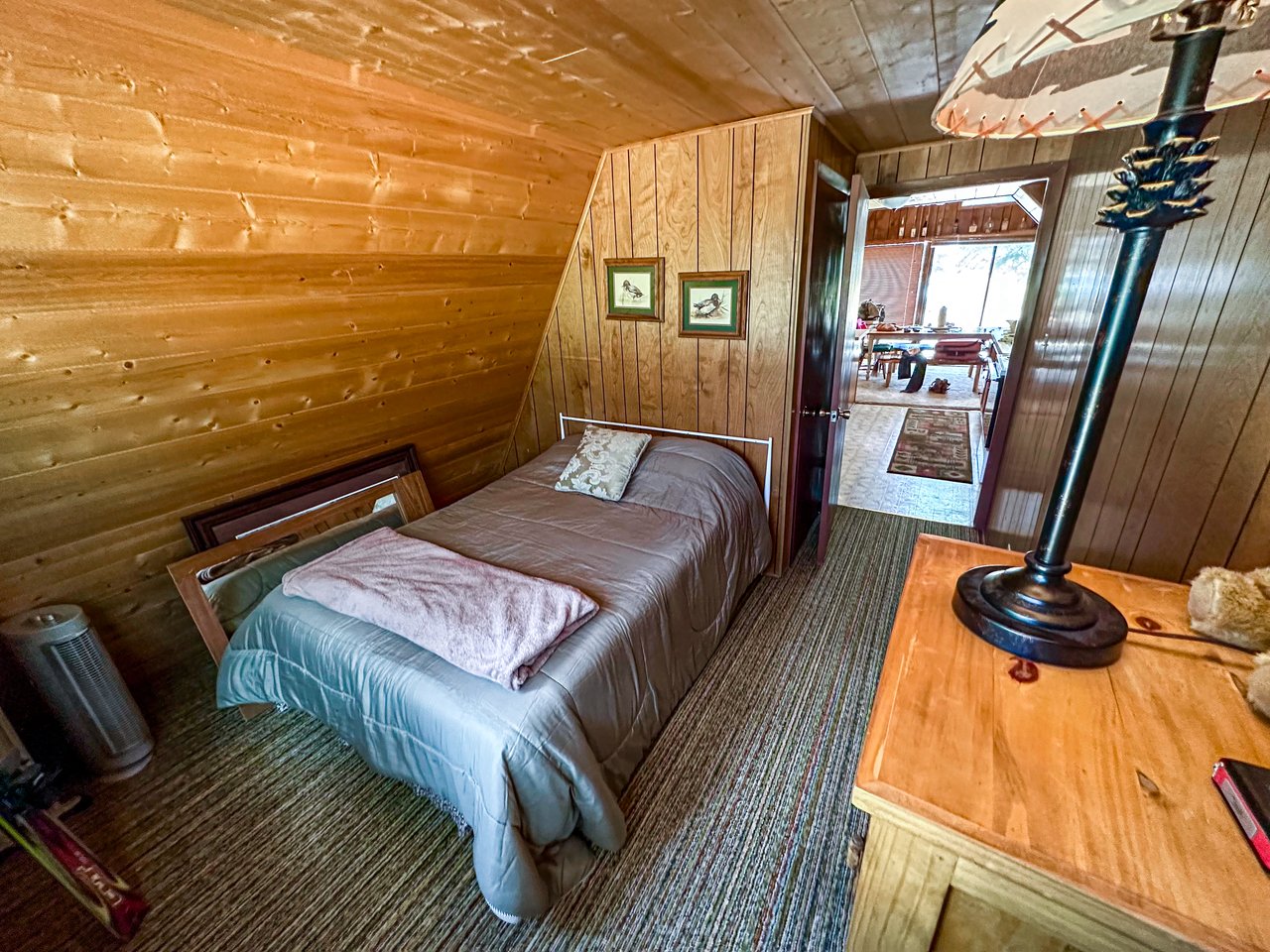 Rustic wood-paneled bedroom with a sloped ceiling, gray-covered bed, and doorway to a dining area.