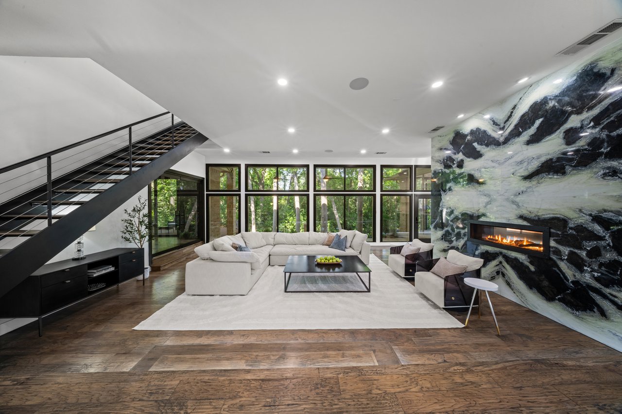 Modern living room with a white sectional, black metal staircase, dramatic stone fireplace, and wooded views.