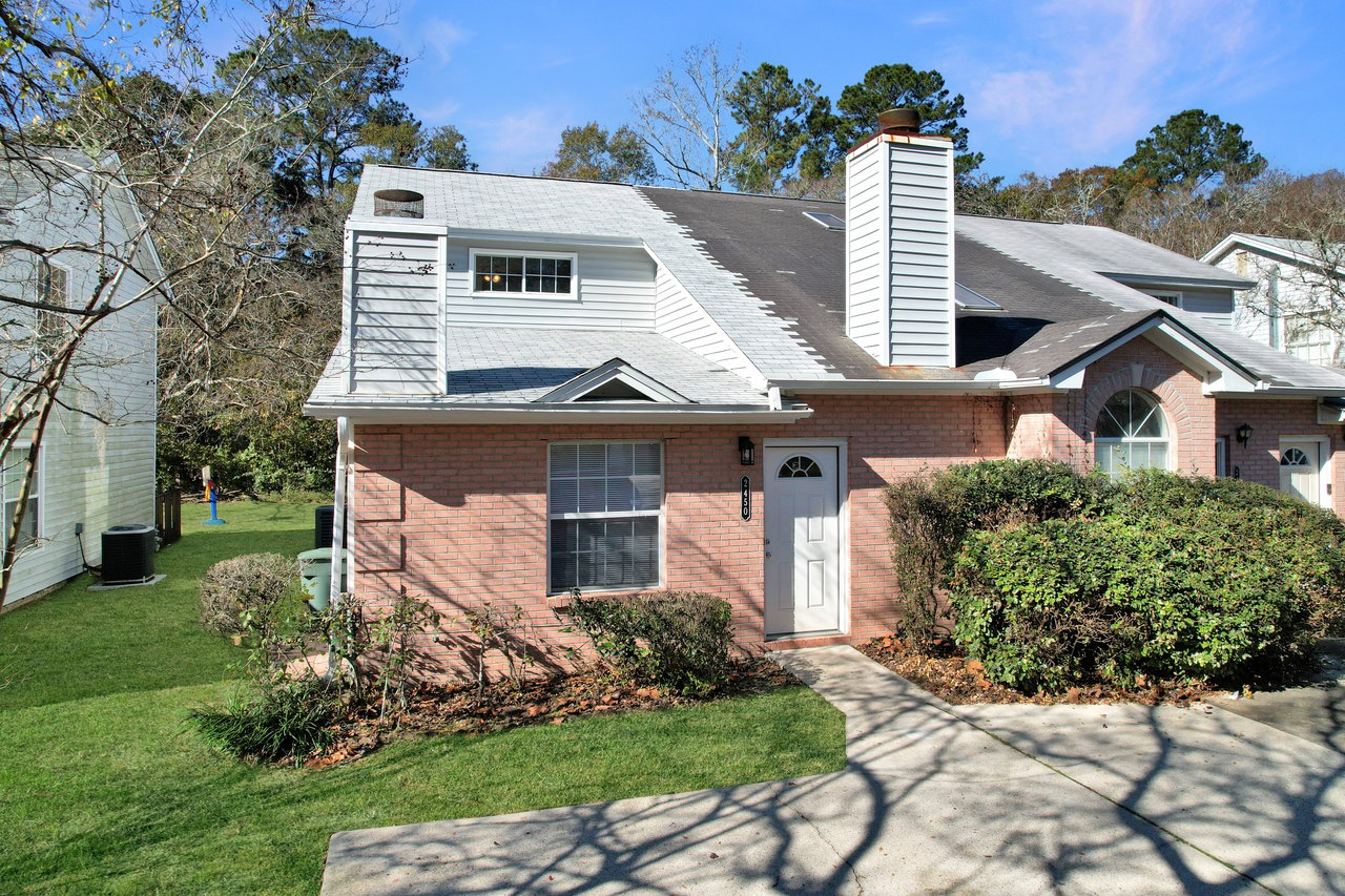 A suburban brick house with white trim, featuring a chimney and large front window. It has a well-maintained lawn, bushes, and a shadowed walkway.