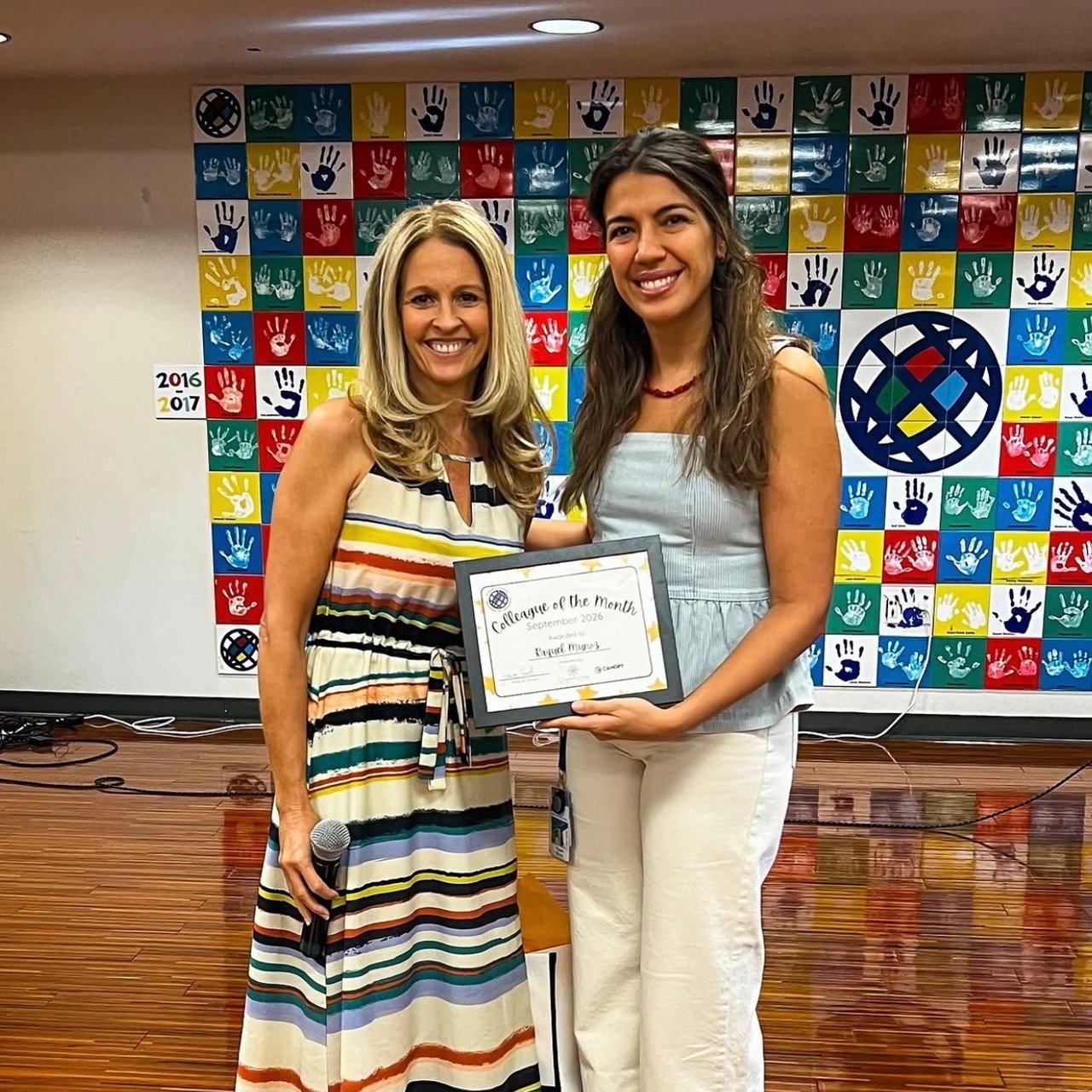 Two women hold a Colleague of the Month certificate in front of a colorful handprint wall.
