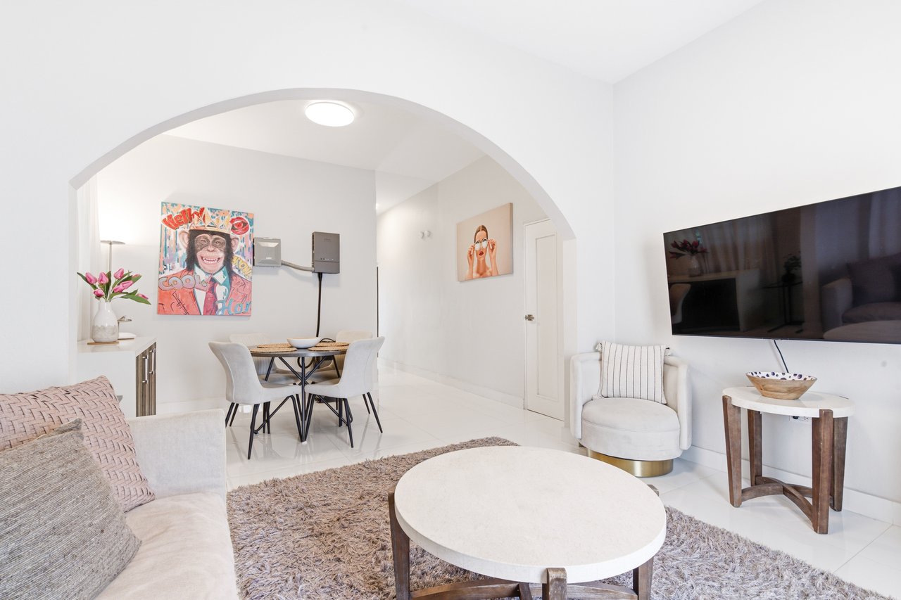 Bright living and dining area with an arched opening, pale furnishings, and white tile floors.