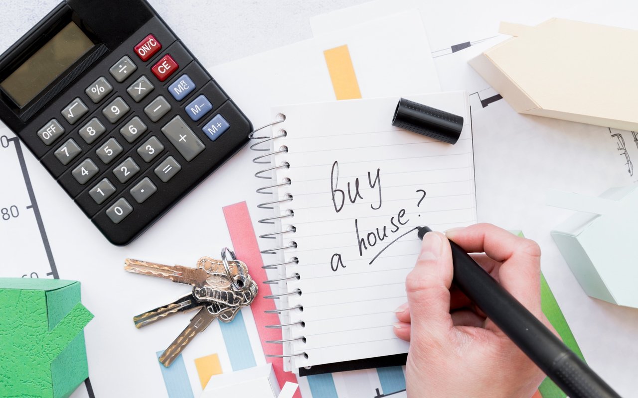 A hand writes “buy a house?” beside a calculator and keys.
