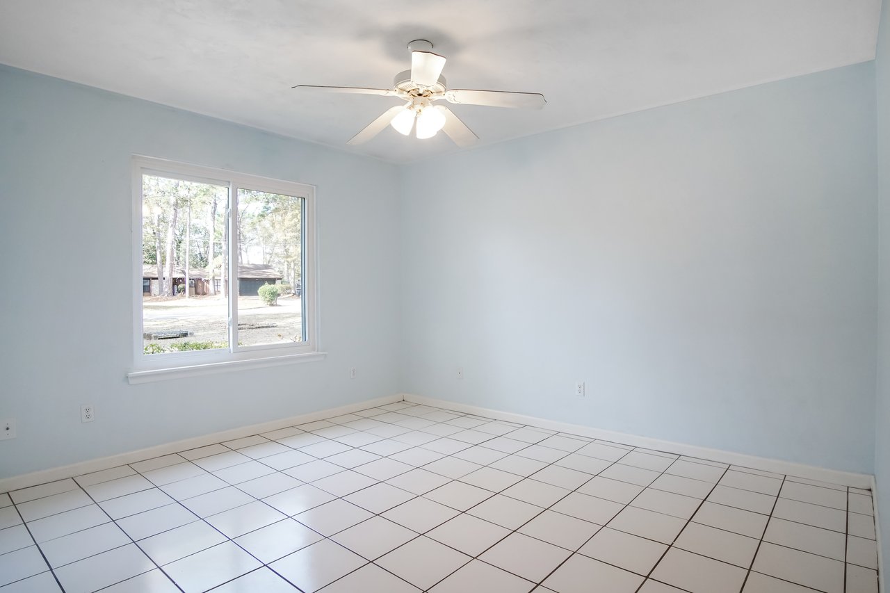 A minimalist room with light blue walls, white tiled floor, and a ceiling fan. A large window offers a view of trees and a house, evoking a serene mood.