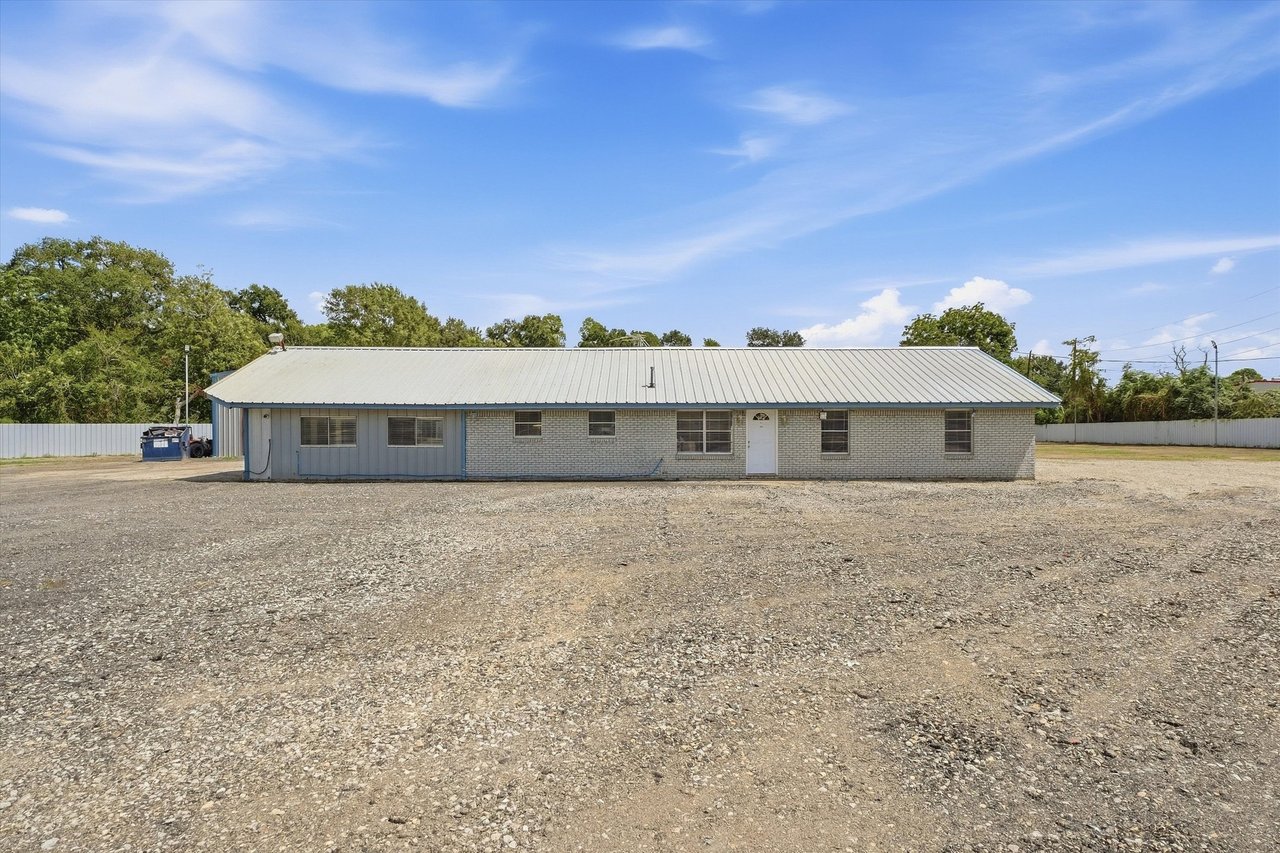 Single-story brick building with a pale metal roof behind a broad gravel lot.