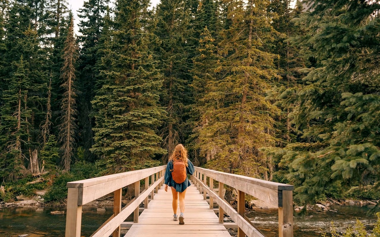 Woman with an orange backpack crossing a wooden bridge through a forest.