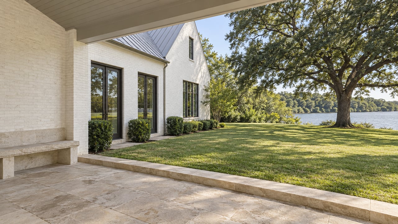 White brick lakeside home with glazed doors, limestone terrace, broad lawn, mature oak, and a narrow strip of water.