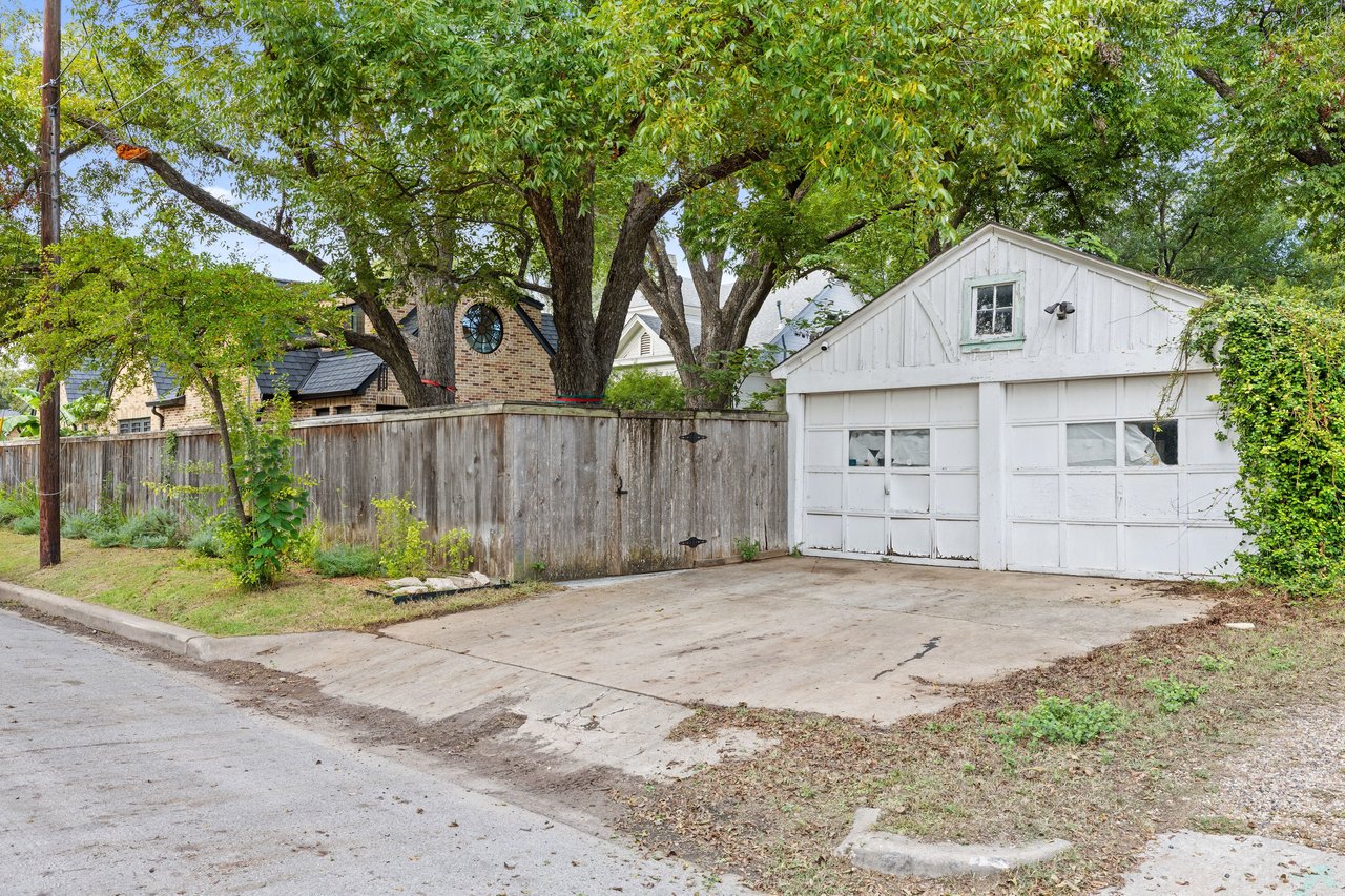 White detached garage beside a concrete driveway, wooden fence, and mature trees.