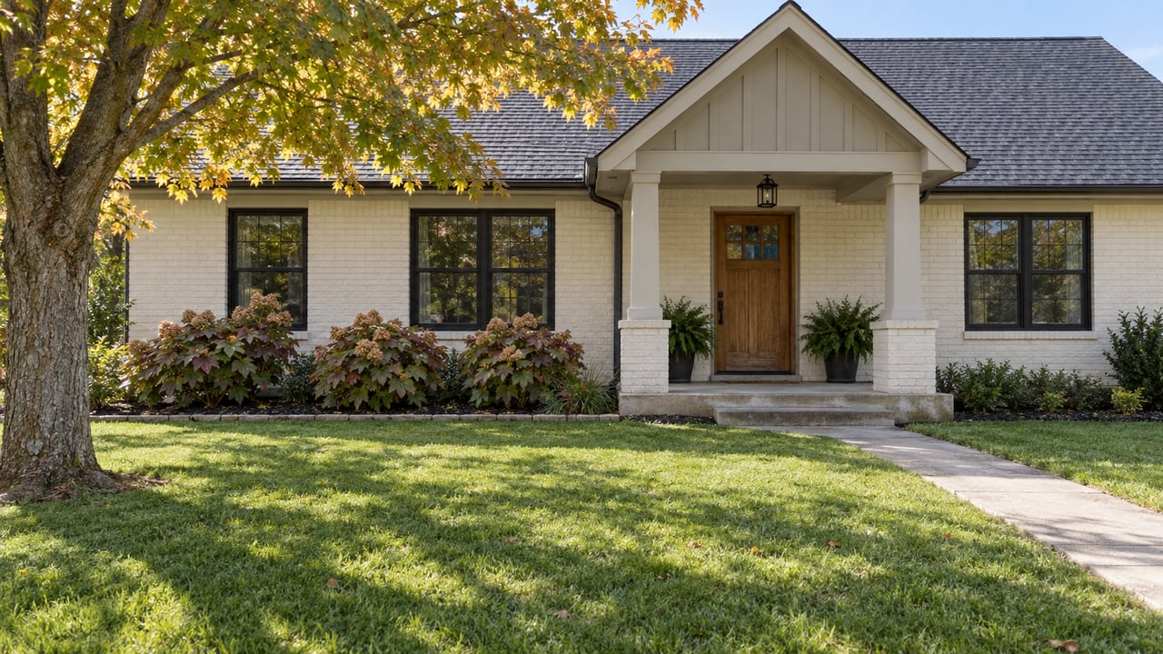 Cream-painted brick ranch with a gabled covered porch, black-framed windows, a broad front lawn, and autumn foliage.